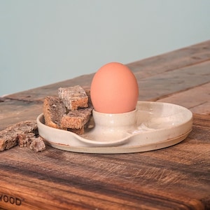 Peut inclure: Un coquetier en céramique avec un œuf brun et des tranches de pain grillé. Le coquetier est blanc cassé et possède une petite coupelle pour le pain. Les éléments sont posés sur une planche à découper en bois.
