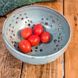 May include: A handmade, light blue ceramic bowl with numerous small holes around the sides. The bowl contains five bright red cherry tomatoes. Dark green kale leaves are visible in the background, resting on a wooden surface. A rustic, handcrafted kitchenware item.