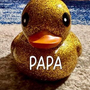 May include: A gold glitter duck with black eyes and an orange beak. The word "PAPA" is written in white block letters across the front. The duck is on a grey surface. A decorative item.