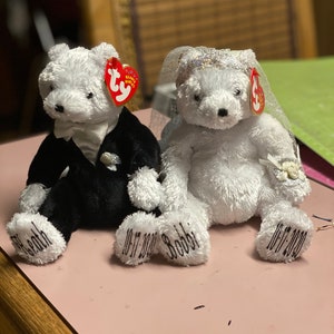 May include: Two white plush teddy bears dressed as a bride and groom. The groom is wearing a black tuxedo and the bride is wearing a white veil. Both bears have the words "10-17-2020" and their names, "Bobby" and "Peach", embroidered on their feet.