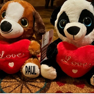 May include: Two plush stuffed animals holding red heart-shaped pillows with the word "Love" embroidered on them. The brown and white dog plush is holding a pillow that says "Love" and has the name "Paul" on its paw. The black and white panda plush is holding a pillow that says "Love" and has the name "Thomas" on its paw.