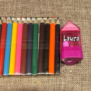 May include: A set of twelve colored pencils in a clear plastic package, next to a pink pencil sharpener. The pencils are in various colors, including red, blue, yellow, and green. The pencil sharpener has the name "Laura" printed on it.