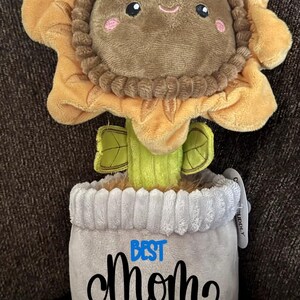 May include: A plush sunflower with a smiling face and a gray pot with the text "BEST MOM" and a red heart.