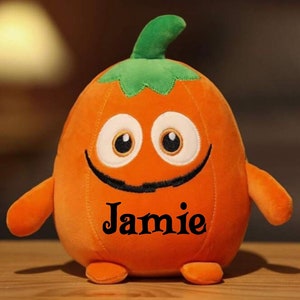 Peut inclure: Citrouille en peluche orange avec un visage souriant, de grands yeux et une moustache noire. La citrouille porte le nom "Jamie" brodé sur le devant.