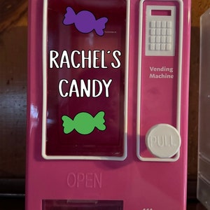 May include: Pink toy vending machine with a white front panel and a clear window. The machine has a pull lever, a coin slot, and a candy dispenser. The window shows two candy pieces, one purple and one green. The machine is labeled "Rachel's Candy" and "Vending Machine".
