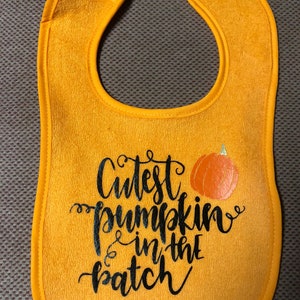 May include: A yellow baby bib with a black and white design that says "Cutest pumpkin in the patch" with a small orange pumpkin graphic.