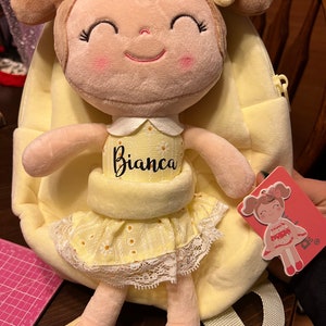 Peut inclure: Un sac à dos en peluche jaune avec une poupée souriante attachée. La poupée a les cheveux bruns, une robe jaune et le nom "Bianca" brodé sur sa poitrine.