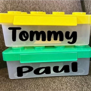 May include: Two rectangular storage containers stacked vertically. The top container has a yellow lid and the name Tommy in black text. The bottom container has a green lid and the name Paul in black text. Both containers are made of clear plastic.