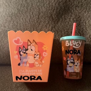 May include: An orange popcorn container and a clear tumbler featuring Bluey characters. The popcorn container has a heart design with the text "I'll always be there for you" and the name "Nora". The tumbler has a blue lid and a pink straw.