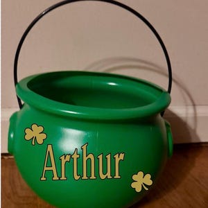 May include: A green plastic pot with a black handle, featuring the name "Arthur" in gold lettering. Two gold shamrock stickers are on the pot. The pot is likely used for St. Patrick's Day.
