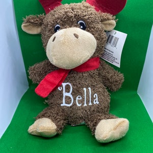 Peut inclure: Un élan en peluche marron avec des bois rouges et une écharpe rouge. L'élan a un nom brodé en blanc "Bella" sur sa poitrine.