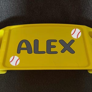 May include: A yellow lap desk with the name "ALEX" in gray block letters. Two baseballs are on the desk, one on each side of the name. The desk has two side compartments and a cup holder.
