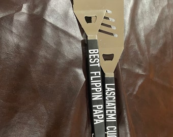 May include: Two black handled grilling spatulas with silver blades. One spatula has the text "Best Flippin Papa" and the other has the text "Lasciatemi Cucinare".