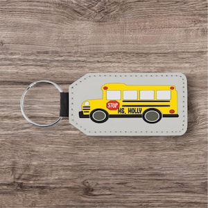 May include: A gray keychain with a yellow school bus design. The bus has black wheels, a red stop sign, and the text "Ms. Holly". The keychain has a silver ring and a black strap.
