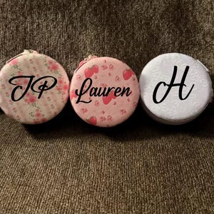 May include: Three round, personalised tape measures. One is floral with "JP", another is pink with strawberries and "Lauren", and the last is white with "H". Each has a small zip.