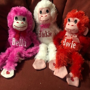 May include: Three plush monkey toys with pink, white, and red fur. Each monkey has a pink ribbon around its neck and a heart-shaped patch on its foot. The monkeys are named Bella, Jackie, and Jovie.