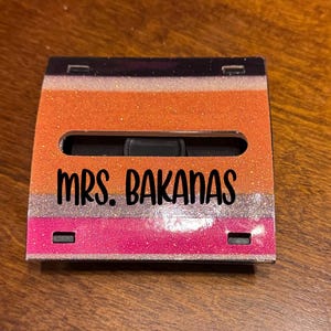 May include: A rectangular case with a glittery design. The top section is black, followed by orange, silver, and pink glitter layers. The text "MRS. BAKANAS" is in black, centered on the case.