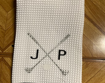 May include: White golf towel with a waffle weave texture. The towel features crossed golf clubs, the initials "J P", and a golf ball graphic. The edges are trimmed with black. A tag at the bottom reads "Made in China".
