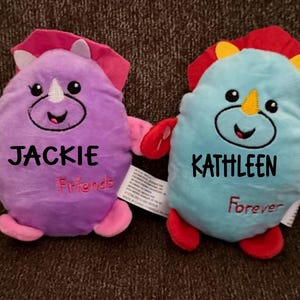 May include: Two plush dinosaur toys, one purple and one blue, each with a smiling face and the name of a person embroidered on the front. The purple dinosaur says "Jackie Friends" and the blue one says "Kathleen Forever".