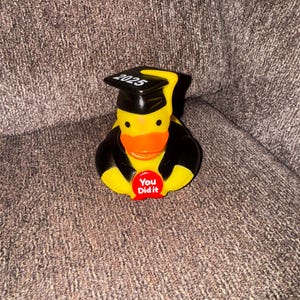 May include: A yellow rubber duck wearing a black graduation cap with "2025" printed on it and a black graduation gown. The duck has an orange beak and a red button that says "You Did it".