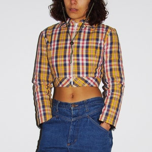 Peut inclure: Une personne portant une chemise courte à carreaux jaune, rouge et bleu avec une fermeture éclair et un jean bleu. Elle porte des bottes de travail orange.