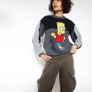 Könnte beinhalten: Ein grauer, schwarzer und roter Pullover mit einem Cartoon-Bild von Bart Simpson beim Skateboardfahren. Der Pullover wird von einer Person getragen, die eine grüne Cargohose und braune Schuhe trägt.