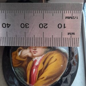 May include: A black and white cameo pendant with a painted portrait of a young person wearing a yellow jacket and red shirt. The cameo is set in a black metal frame with a scalloped edge. The pendant is measured against a ruler, showing it is approximately 2.5 inches in diameter.