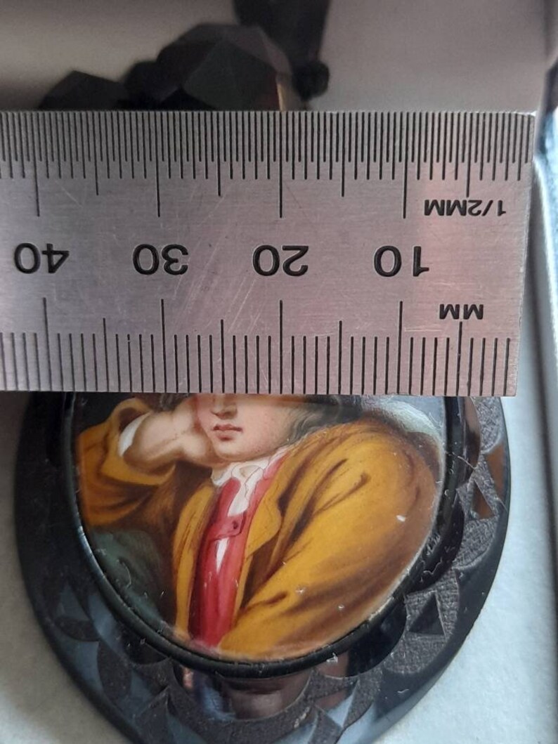 May include: A black and white cameo pendant with a painted portrait of a young person wearing a yellow jacket and red shirt. The cameo is set in a black metal frame with a scalloped edge. The pendant is measured against a ruler, showing it is approximately 2.5 inches in diameter.