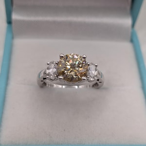 May include: A three-stone ring featuring a large, round, champagne-coloured centre gemstone flanked by two smaller, oval-shaped, clear white gemstones. The stones are set in a polished silver-toned metal band with four prongs for the centre stone.