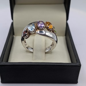 May include: A silver ring featuring a curved design, adorned with a selection of colourful gemstones. The gemstones are red, blue, purple, yellow, and green. The ring is presented in a black jewellery box with a cream-coloured interior.