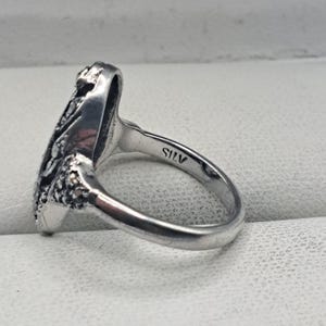 May include: A silver ring with an ornate design on the face. The ring has a smooth band and a detailed, textured pattern on the top. The word "SILV" is stamped inside the band. The ring is likely made of sterling silver.