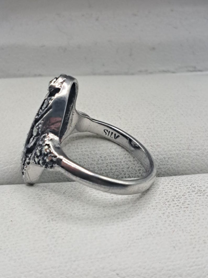May include: A silver ring with an ornate design on the face. The ring has a smooth band and a detailed, textured pattern on the top. The word "SILV" is stamped inside the band. The ring is likely made of sterling silver.