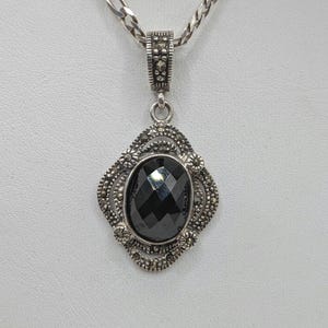 Op de afbeelding: Zilveren hangerketting met een zwarte, ovaal geslepen edelsteen. De edelsteen is gezet in een decoratief montuur van markasiet. De ketting heeft een zilveren ketting.