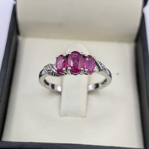 May include: A silver ring featuring three oval-cut, vivid red gemstones. The ring is set in a silver band with small accent stones. The ring is presented in a black jewellery box with a white interior.