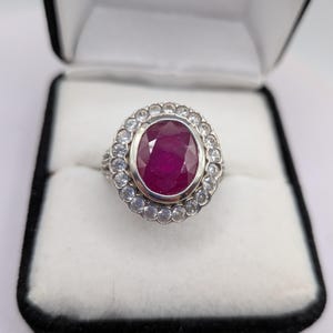 May include: A silver-toned ring featuring a large, oval-cut deep red gemstone at the centre, surrounded by a halo of small, clear, round-cut stones. The band is metallic and the ring is presented in a black and white jewellery display box.