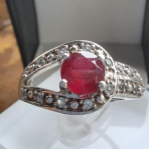 Puede incluir: Un anillo de plata con una gran piedra preciosa de rubí rojo rodeada de piedras blancas más pequeñas.
