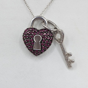 May include: A silver necklace with a heart-shaped pendant, set with dark red gemstones, and a key charm. The heart pendant has a keyhole design. The necklace is displayed against a white backdrop. The key and pendant are detailed.