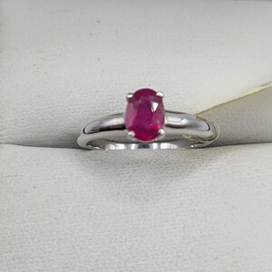 May include: A silver ring featuring a vibrant oval-cut ruby gemstone. The ruby is a deep, rich red color, set in a classic prong setting. The band is a polished silver, creating a simple yet elegant design.
