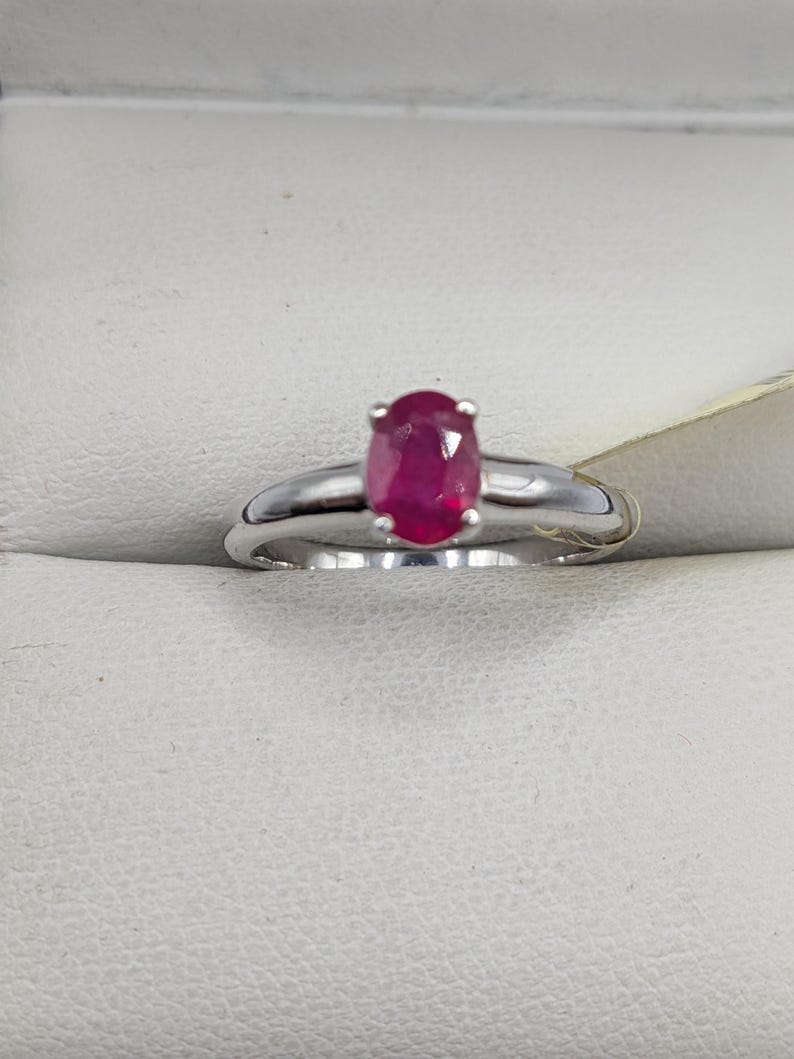 May include: A silver ring featuring a vibrant oval-cut ruby gemstone. The ruby is a deep, rich red color, set in a classic prong setting. The band is a polished silver, creating a simple yet elegant design.