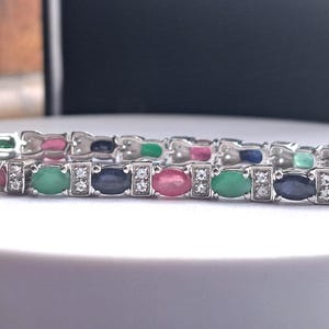 Puede incluir: Una pulsera de plata con piedras preciosas ovales de varios colores, incluyendo rosa, verde y azul. Pequeñas piedras transparentes se intercalan entre las gemas más grandes. La pulsera se muestra sobre una superficie blanca.