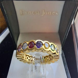 May include: A gold band ring with a row of colorful gemstones. The ring is in a black box with the words "Ernest Jones" printed on the inside.
