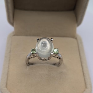 May include: A silver ring with a large, oval, translucent gemstone at its centre, with two smaller, round, green gemstones either side. The ring is set in a silver band and displayed in a beige ring box.