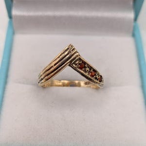 Op de afbeelding: Een V-vormige gouden ring met een getextureerd, geribbeld ontwerp aan de ene kant en een rij van drie kleine, ronde, donkerrode edelstenen aan de andere kant. De ring heeft een gepolijste afwerking en een puntig chevron-silhouet.