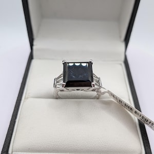 May include: A silver ring with a large, rectangular, dark gemstone is displayed in a black box with a white interior. The ring has smaller, clear stones on either side of the main stone. The box is labeled "ERNEST JONES" on the inside of the lid.