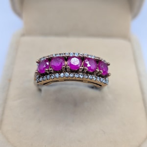 Può includere: Un anello con cinque pietre rotonde di un rosa vivace disposte in fila, affiancate da due file di piccole pietre bianche trasparenti. La fascia è realizzata in metallo scuro con finitura anticata, creando un contrasto con le pietre.
