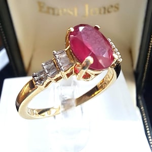 Può includere: Un anello d'oro con un grande rubino ovale e pietre rettangolari e di accento più piccole. L'anello è presentato in una scatola nera con la scritta "Ernest Jones". Il rubino è il punto focale dell'anello.