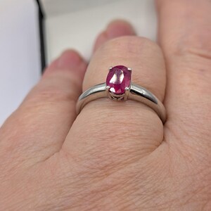 May include: A silver ring featuring a vibrant oval-cut ruby gemstone. The ruby is a deep red color and is set in a simple, elegant band. The ring is shown on a finger.