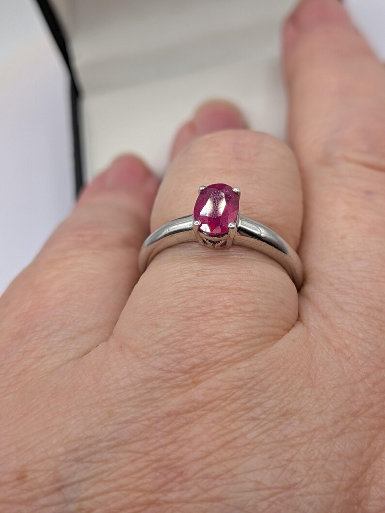 May include: A silver ring featuring a vibrant oval-cut ruby gemstone. The ruby is a deep red color and is set in a simple, elegant band. The ring is shown on a finger.
