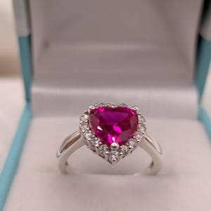 May include: A silver ring featuring a large, heart-shaped, vibrant pink gemstone as the centrepiece. The central stone is surrounded by a halo of small, clear, round-cut white stones set into the silver band, creating a bright and reflective appearance.