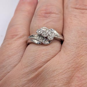 May include: A silver ring with a cluster of small round diamonds and baguette-cut diamonds. The ring has a swirling design with the diamonds set in a raised setting. The ring is on a finger.